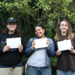 Three students from The King Alfred School smile and hold their A level exam results certificates in front of a bush