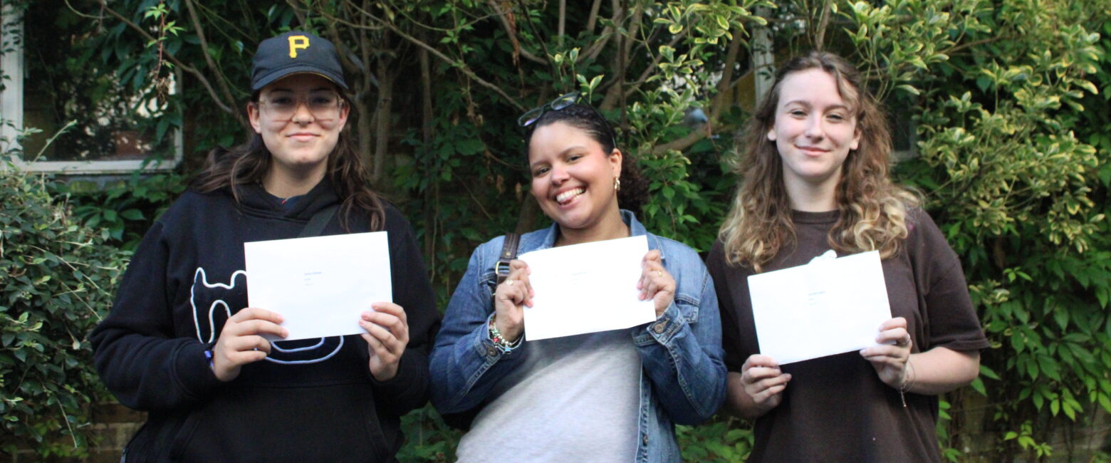 Three students from The King Alfred School smile and hold their A level exam results certificates in front of a bush