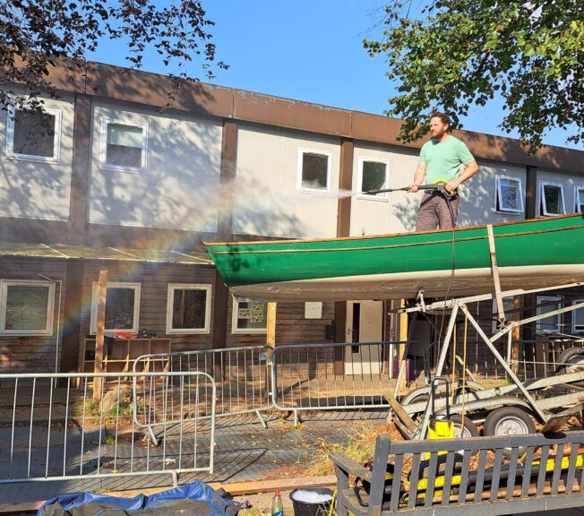 A man stands in a green boat atop a trailer spraying a hose, creating a rainbow