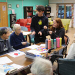 Sixth Form students from The King Alfred School and care home residents sit around a table with tubes of paint and paper