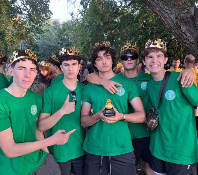 Five male students from The King Alfred School Sixth Form wear green school t-shirts and comedy crowns, with one holding a rubber duck trophy