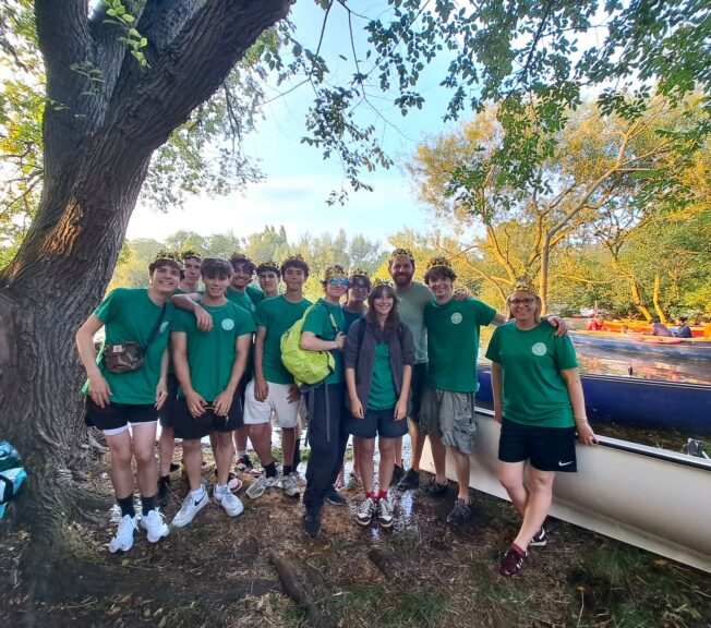 A group of ten students and two teachers pose on the banks of the River Thames alongside a rowing boat