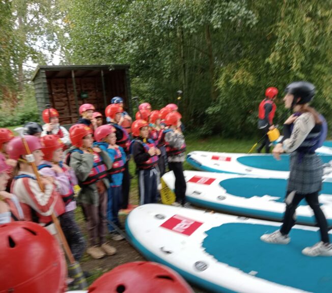 Students from The King Alfred School stand in a group whilst a paddleboarding instructor shows them how to use the boards