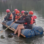 Students from The King Alfred School sit atop a self-made raft on a lake, whilst a teacher uses a paddle to row