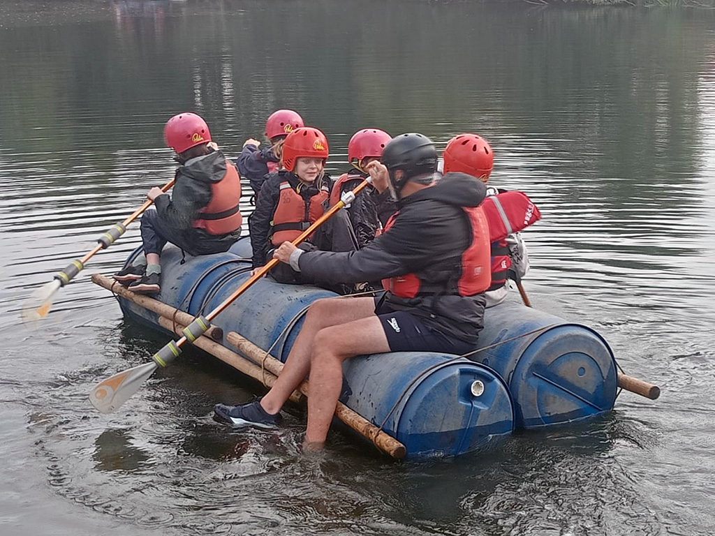 Students from The King Alfred School sit atop a self-made raft on a lake, whilst a teacher uses a paddle to row