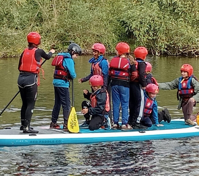 Eight students from The King Alfred School stand on a paddleboard on a lake