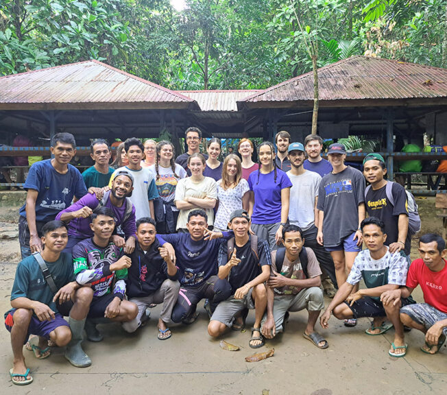 A group of men, women and teens, made up of staff and students from The King Alfred School and Indonesian conservation workers, pictured in front of rainforest huts and trees