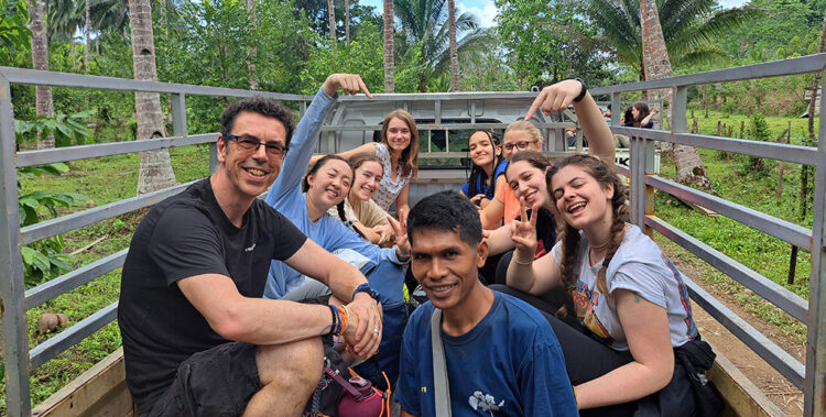 A group of students and staff members from The King Alfred School sit smiling in the back of an open-air truck with palm trees in the background