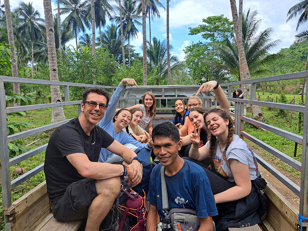 A group of students and staff members from The King Alfred School sit smiling in the back of an open-air truck with palm trees in the background