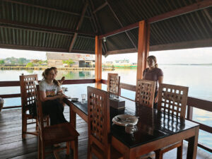 Three teenage girls sit around a wooden table in a open-sided hut on a river in Indonesia