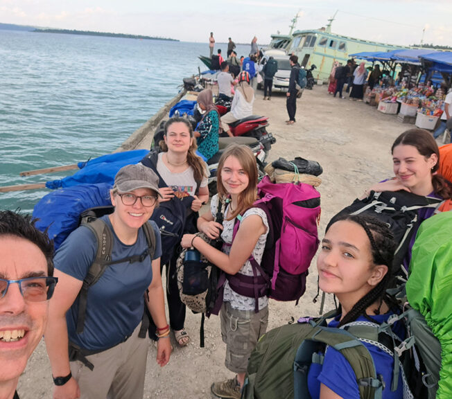 Staff and students from The King Alfred School stand on a jetty next to the sea in Indonesia with a boat and market in the background