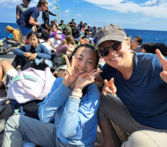 A staff member from The King Alfred School make peace signs and smiles with a student whilst sitting on a crowded boat on the sea in Indonesia