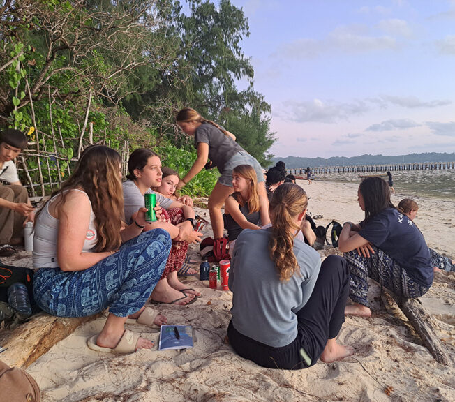 A group of students from The King Alfred School sit on a beach at dusk drinking soft drinks