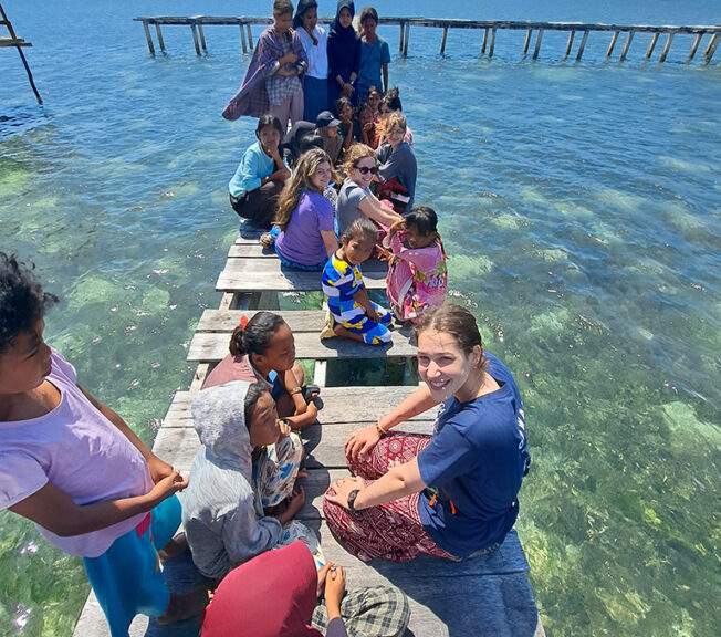 A student from The King Alfred School kneels on a jetty above the sea in Indonesia and smiles at the camera, with local children around her