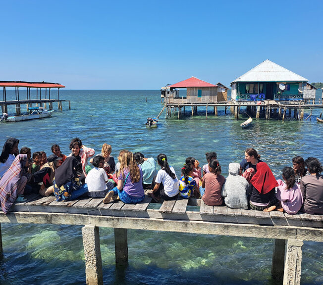 Students from The King Alfred School sit on a wooden jetty over the sea with local Indonesian children, with sea huts in the background