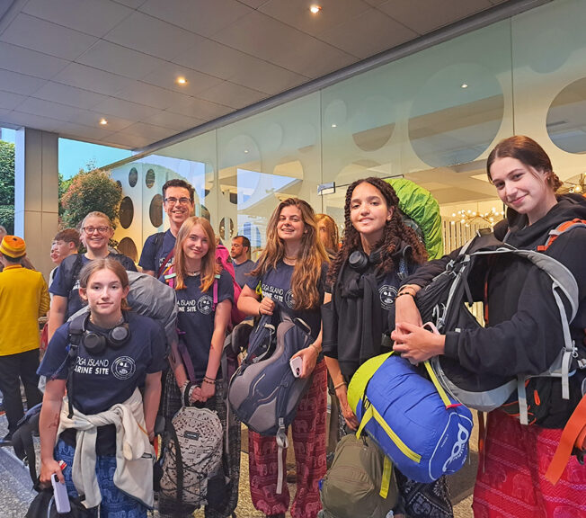 A group of staff and students from The King Alfred School dressed in casual clothing and carrying big backpacks smile in front of an airport terminal