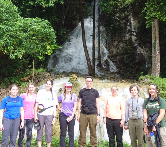 A group of staff and students from The King Alfred School stand in a line in front of a waterfall in Indonesia