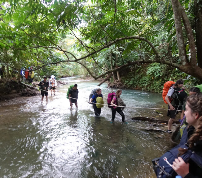 A group of students from The King Alfred School cross a river in the Indonesian rainforest whilst holding onto a rope