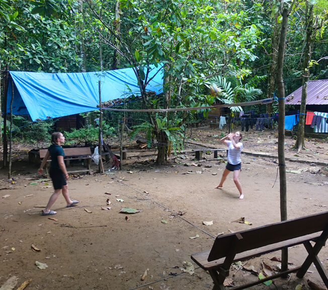 Two students from The King Alfred School play volleyball on a mud court in the Indonesian rainforest, with huts and washing hanging behind them