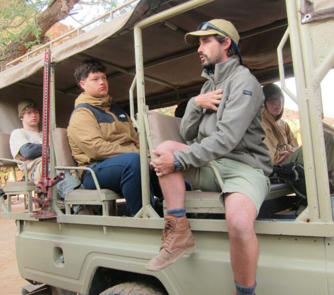 A tour guide sits with his legs over the side of an open-back truck, talking to students sat in the back