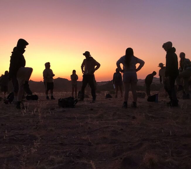 Students from The King Alfred School in silhouette standing in the Namibian desert as the sun sets