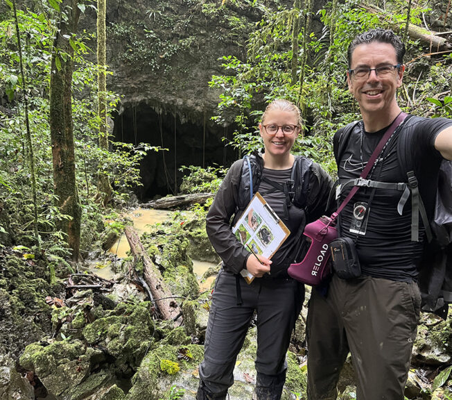 A male and female member of staff from The King Alfred School smile in front of a cave in the Indonesian rainforest, holding a clipboard, camera, compas and water bottle