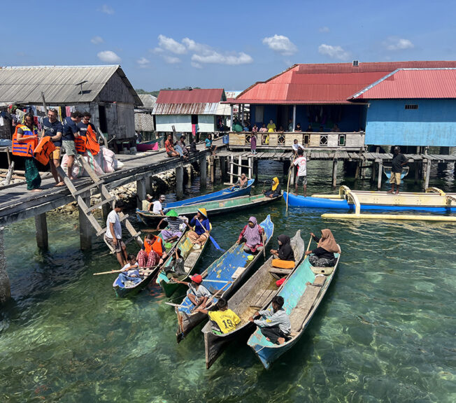 Narrow, colourful boats pictured in the water next to a jetty in Indonesia, with people boarding down a wooden ladder