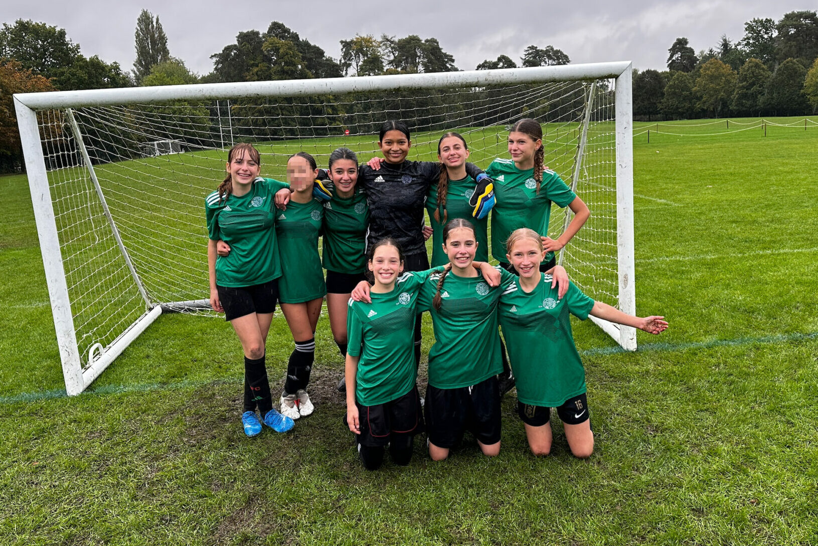 Year 8 girls from The King Alfred School pose in front of a football goal, wearing green jerseys. Five are standing and three kneel in front.