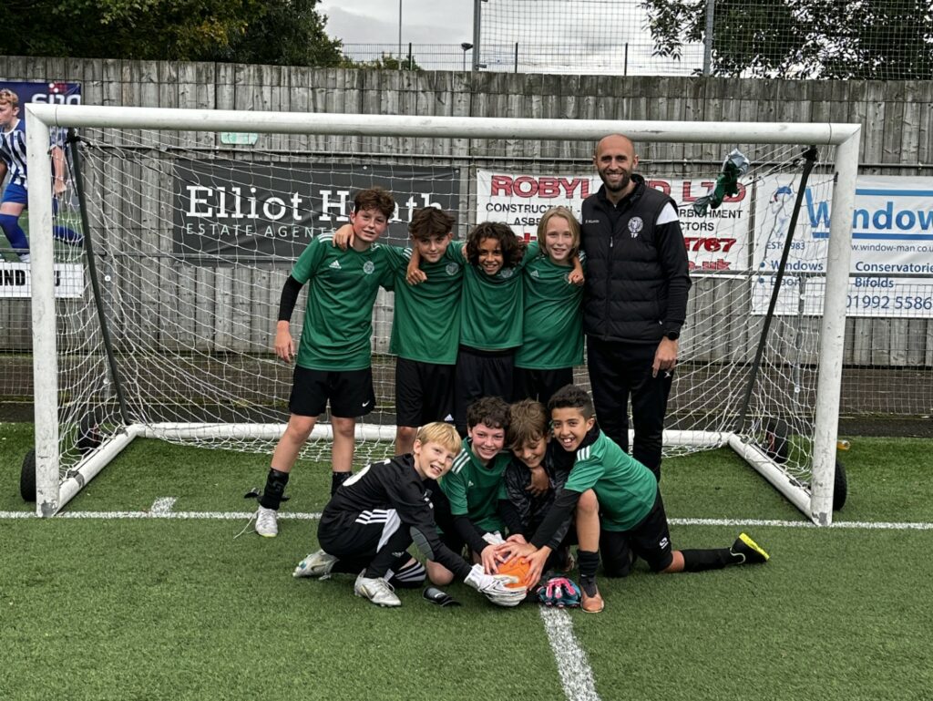 Year 7 boys from The King Alfred School pose with their male coach in front of a football goal. They wear green jerseys and black shorts, with the goalkeeper and coach in all black. Five stand and four kneel in front.