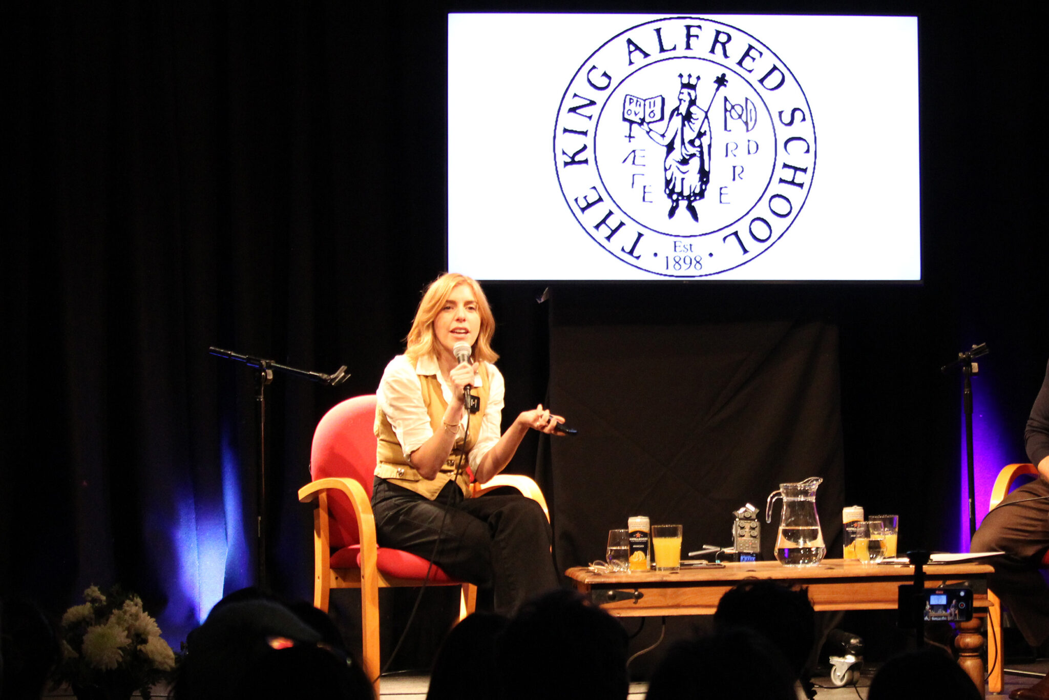 A woman holds a microphone whilst sitting on a stage at The King Alfred School