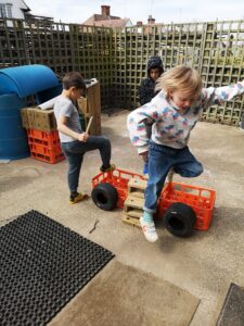 Three young boys play outdoors. They are jumping over a 'car' they have made out of milk crates and old typres.