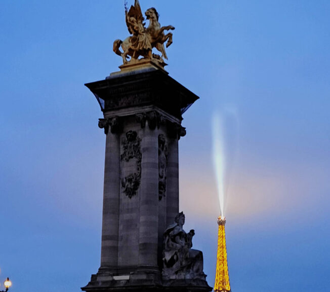 Gold statue on a stone monument with the Eiffel Tower in the background lit up against a dark blue sky