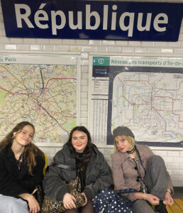 Three female Sixth Form students from The King Alfred School sit on a bench on the platform of a Paris Metro station. There is a map on the wall behind them and the word 'Republique' in large letters on a sign.