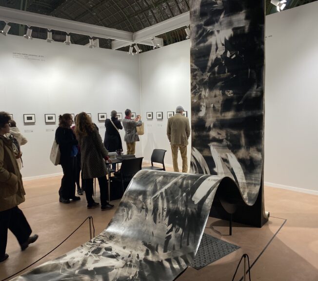 A giant reem of 'paper' featuring black and white pictures stands in the middle of a room at Paris Photo, with people around looking at it and other photos on the walls