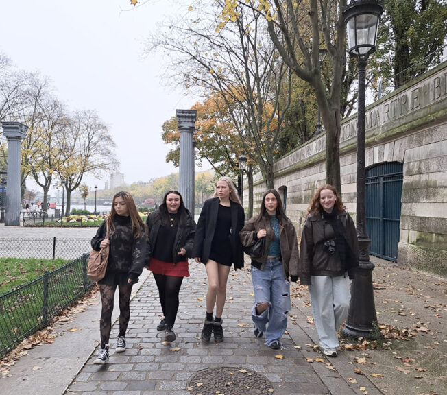 Five female Sixth Form students from The King Alfred School walk in a line along a street in Paris.