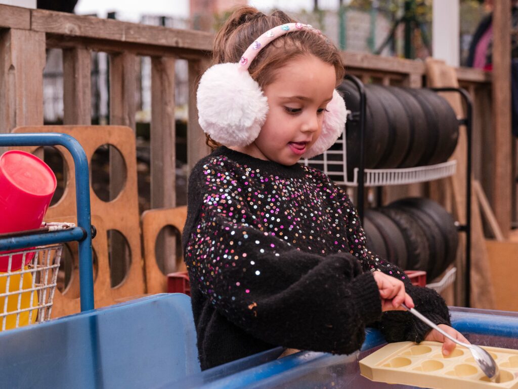 Young girl enjoys loose parts play with a spoon and ice cube tray outside. She wears a black jumper and earmuffs, and items including tyres, a bucker and wooden pallets are in the background.