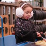 Young girl enjoys loose parts play with a spoon and ice cube tray outside. She wears a black jumper and earmuffs, and items including tyres, a bucker and wooden pallets are in the background.