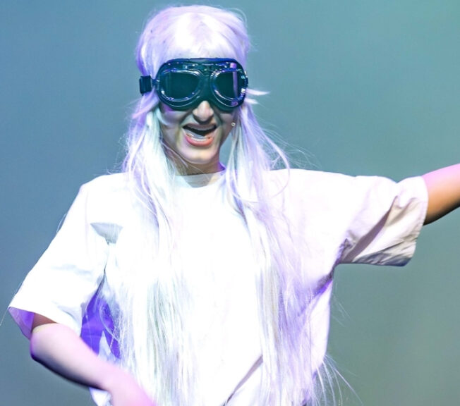 A performer dressed as a scientist in a white lab coat and goggles, holding a skull