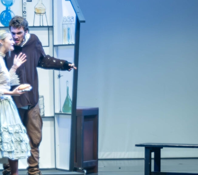 Actors on stage. One is dressed in a black hooded cape, whilst the others look scared