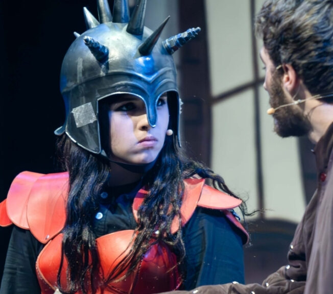 A female actor, wearing a spiky helmet, glowers at another