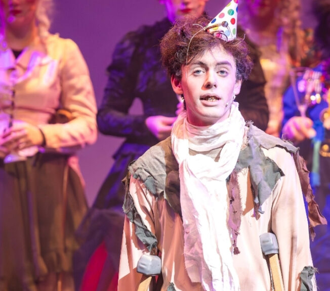 An actor wears a party hat and walks with crutches on stage