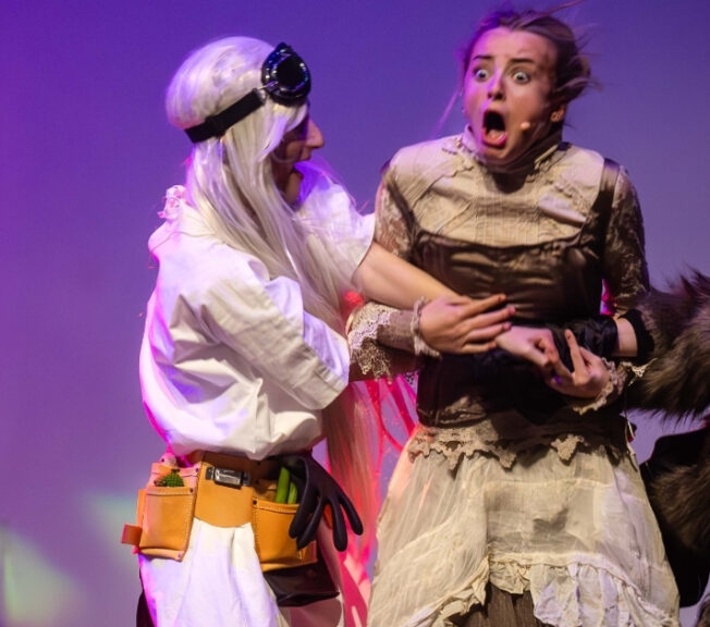 A blonde actor looks surprised as she is seized by fellow performers