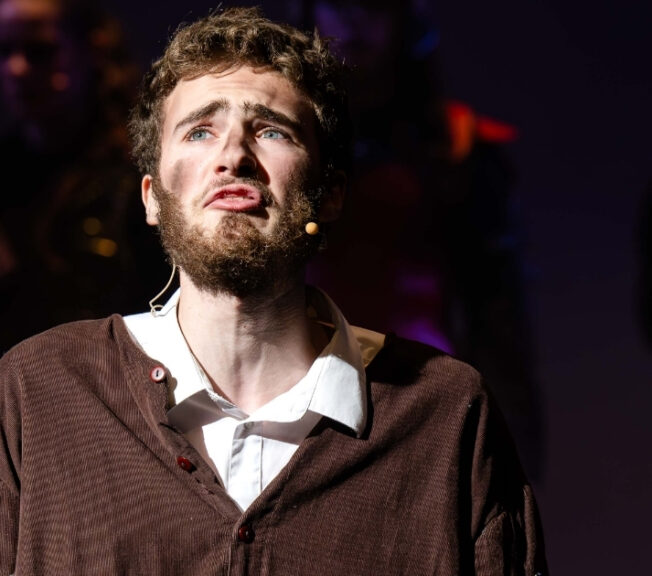 A beadered male actor in a brown smock over a white shirt, with dirt on his face