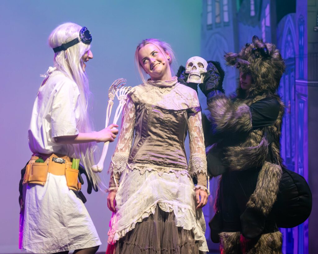 Three actors on stage – a girl in the middle, flanked by a scientist on one side and a giant spider on the other