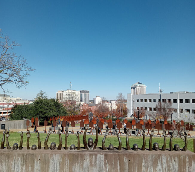 A line of sculptures depicting African Americans emerging from a concrete wall, with a park and blue sky in the background