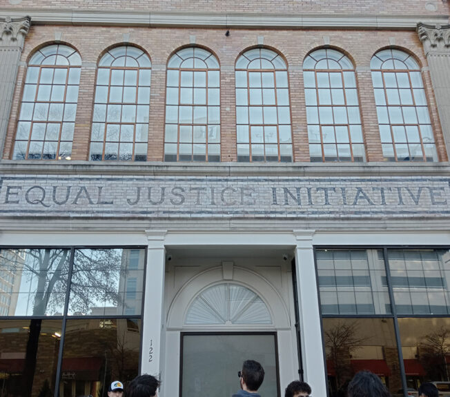 Students stand in front of a stone building with arch windows and the words 'Equal Justice Initiative' spelled out in mosaic