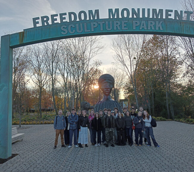 Students from The King Alfred School Sixth Form stand in a park, underneath an arch with the words 'Freedom Monument' on it