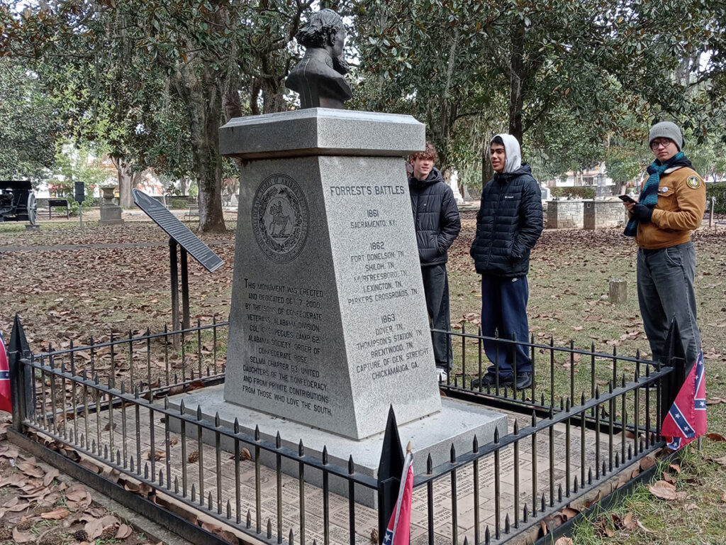 Students consider as tone monument topped with a bust of Nathan Bedford Forrest, surrounded by railings and Confederate flags in a cemetery