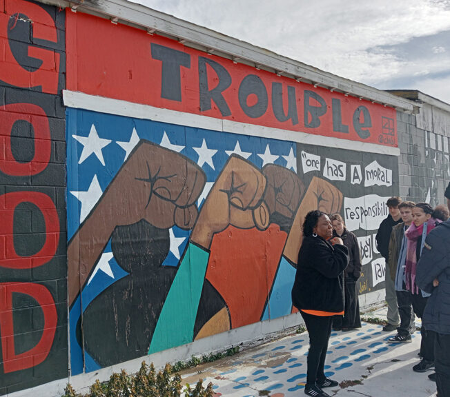 A woman speaks to students in front of a mural that depicts black people raising their fists, and reads 'Good Trouble: one has a moral duty to disobey unjust laws'