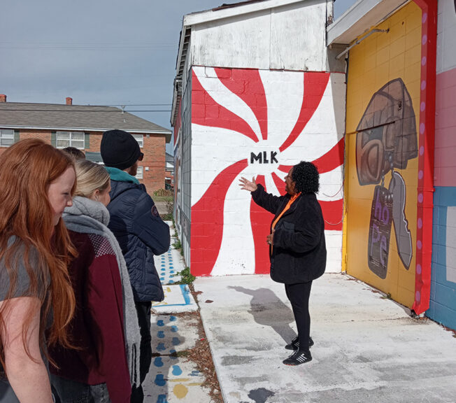 A woman speaks to students in front of a mural with the initials 'MLK' in the centre